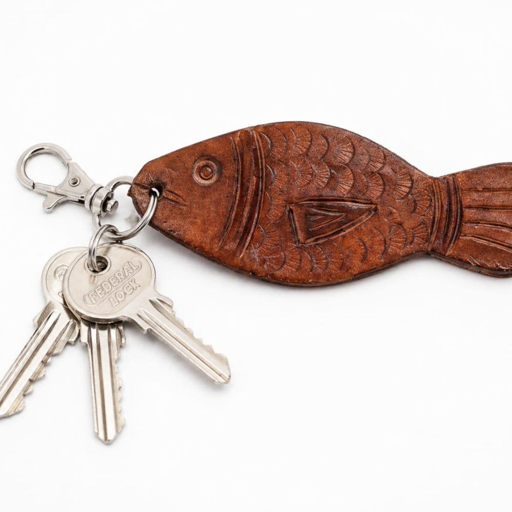 A tan leather fish key fob with tooled scales, clipped to a metal ring with three keys, on a plain pale background.