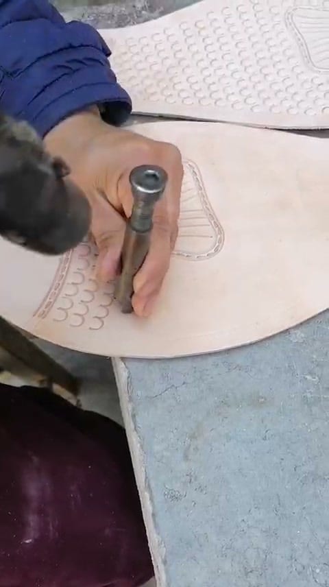 A close view of an artisan's hand holding a steel punch against a pale leather panel, pressing rows of small half-circles into the hide; a finished scaled panel lies on the bench behind.